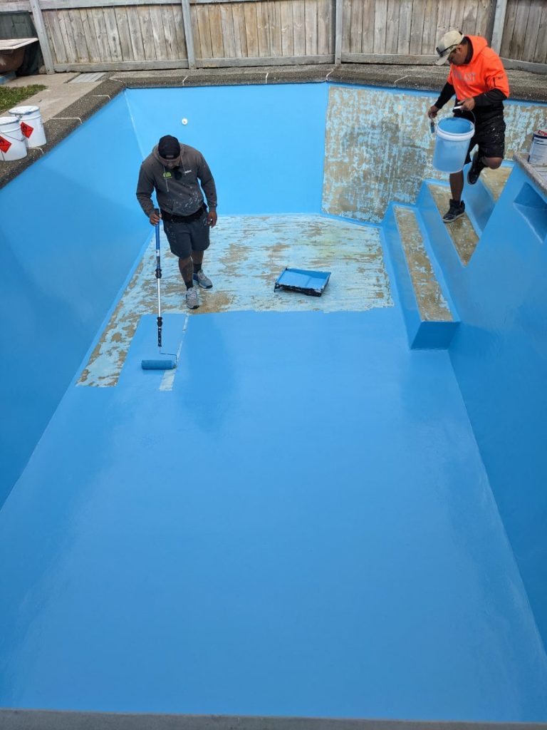 Two painters applying blue pool paint to the interior of a concrete swimming pool in New Zealand